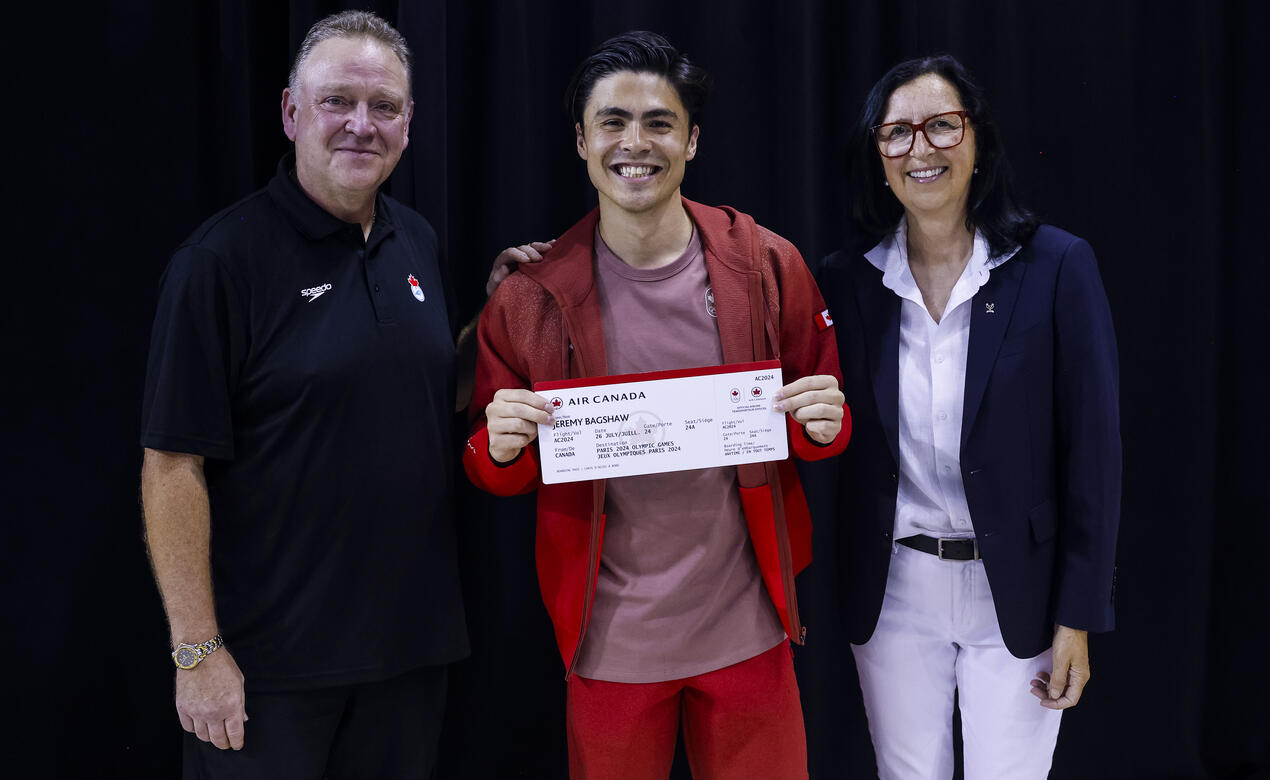 SMUS Alum Jeremy Bagshaw stands centre in full Olympic branded clothing with an enlarged Air Canada plane ticket to Paris 2024. Jeremy stands between two Team Canada leadership members.