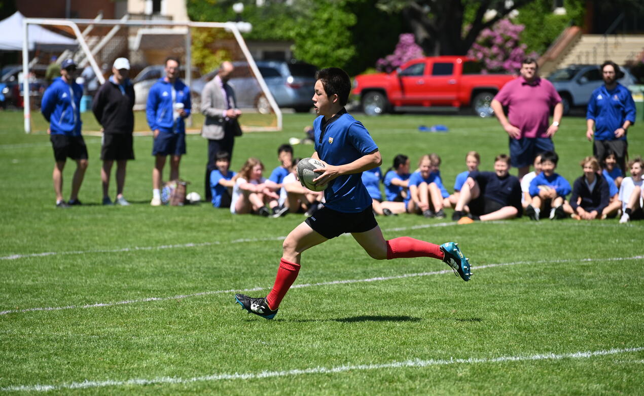 SMUS Junior Boys rugby player runs down the field with the ball as SMUS supporters look on from the sidelines