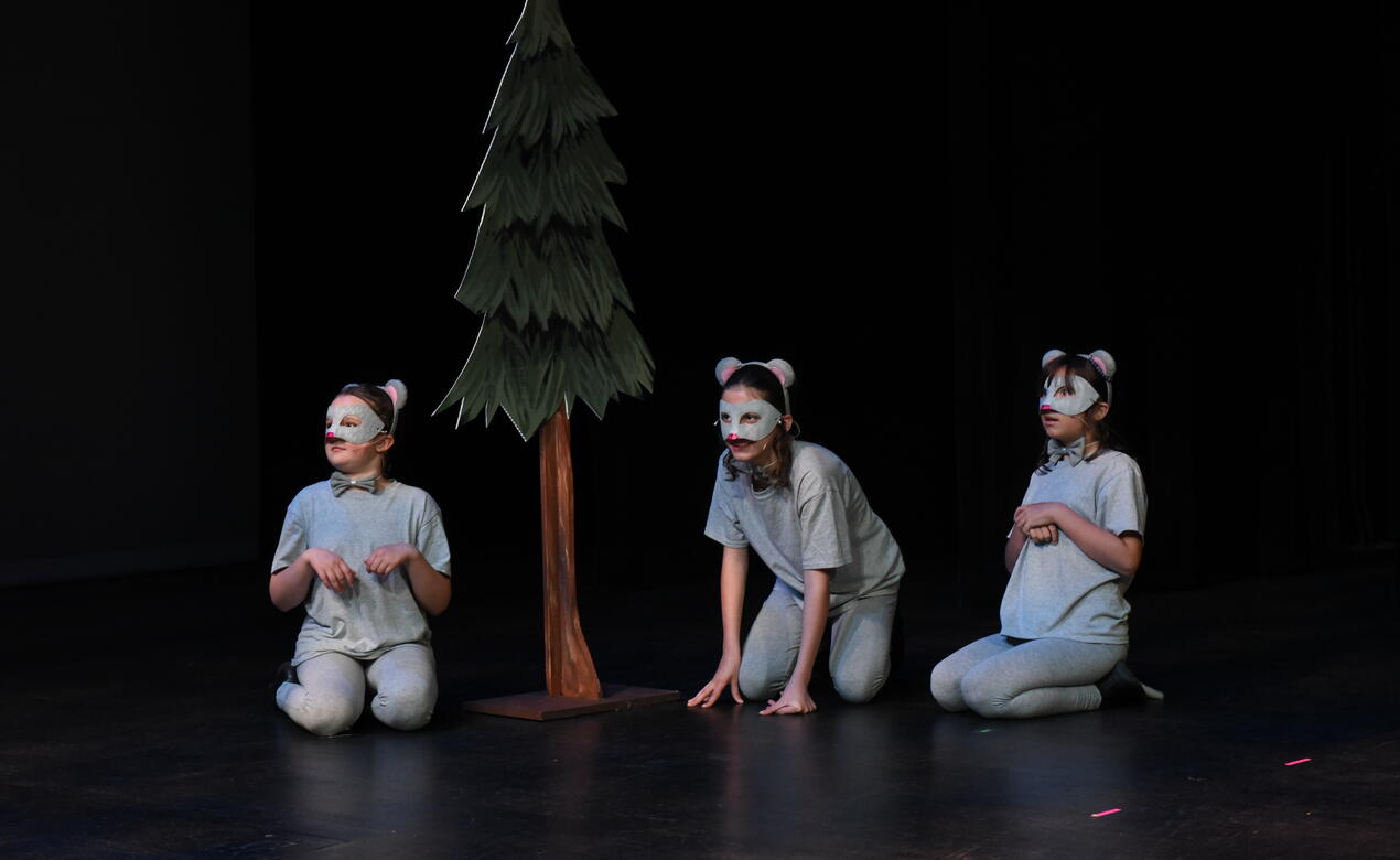 Three students dressed as wolves sit on their knees by a lone tree on stage.