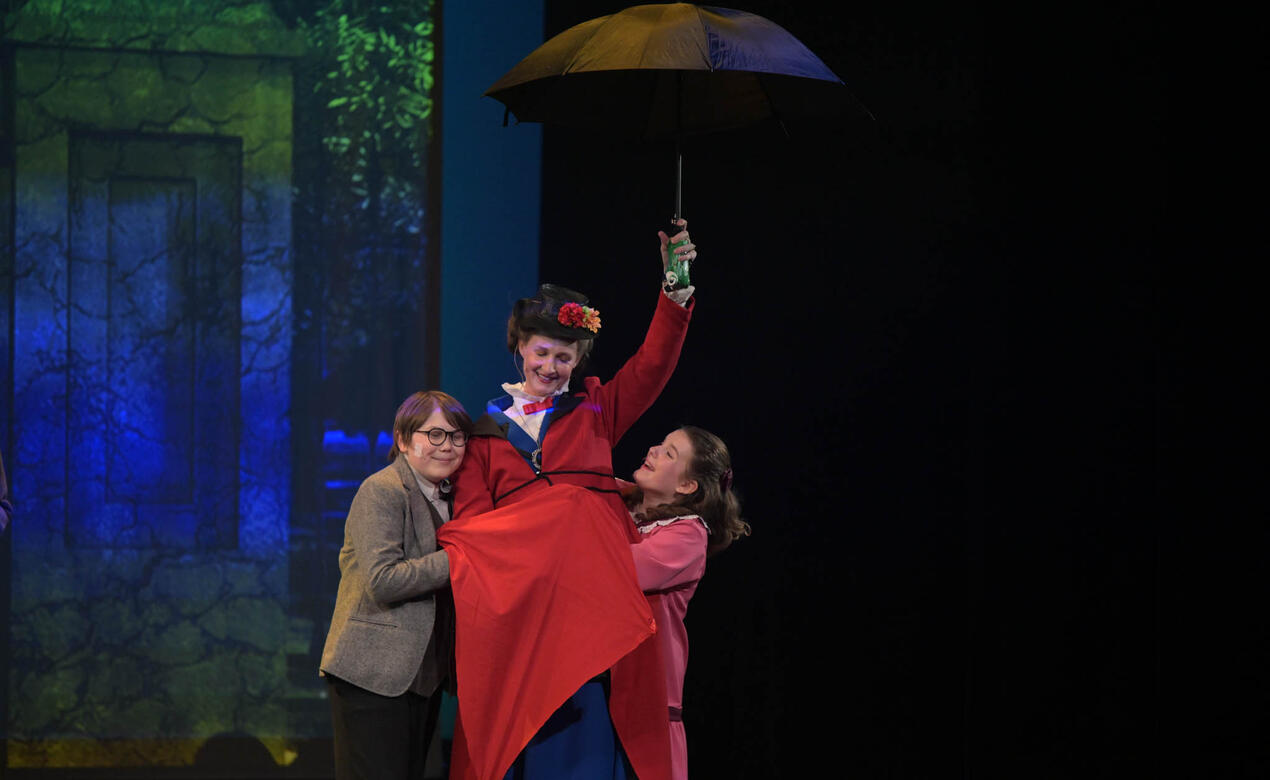 Levi Budd and another cast member hold up Mary Poppins in a red dress with her umbrella open above her.