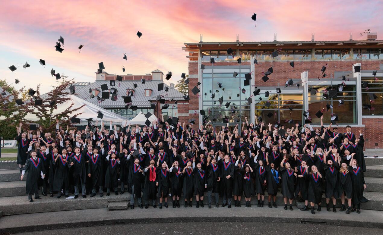 The Graduating Class of 2024 toss their caps in the air as a twilight sky passes over them.