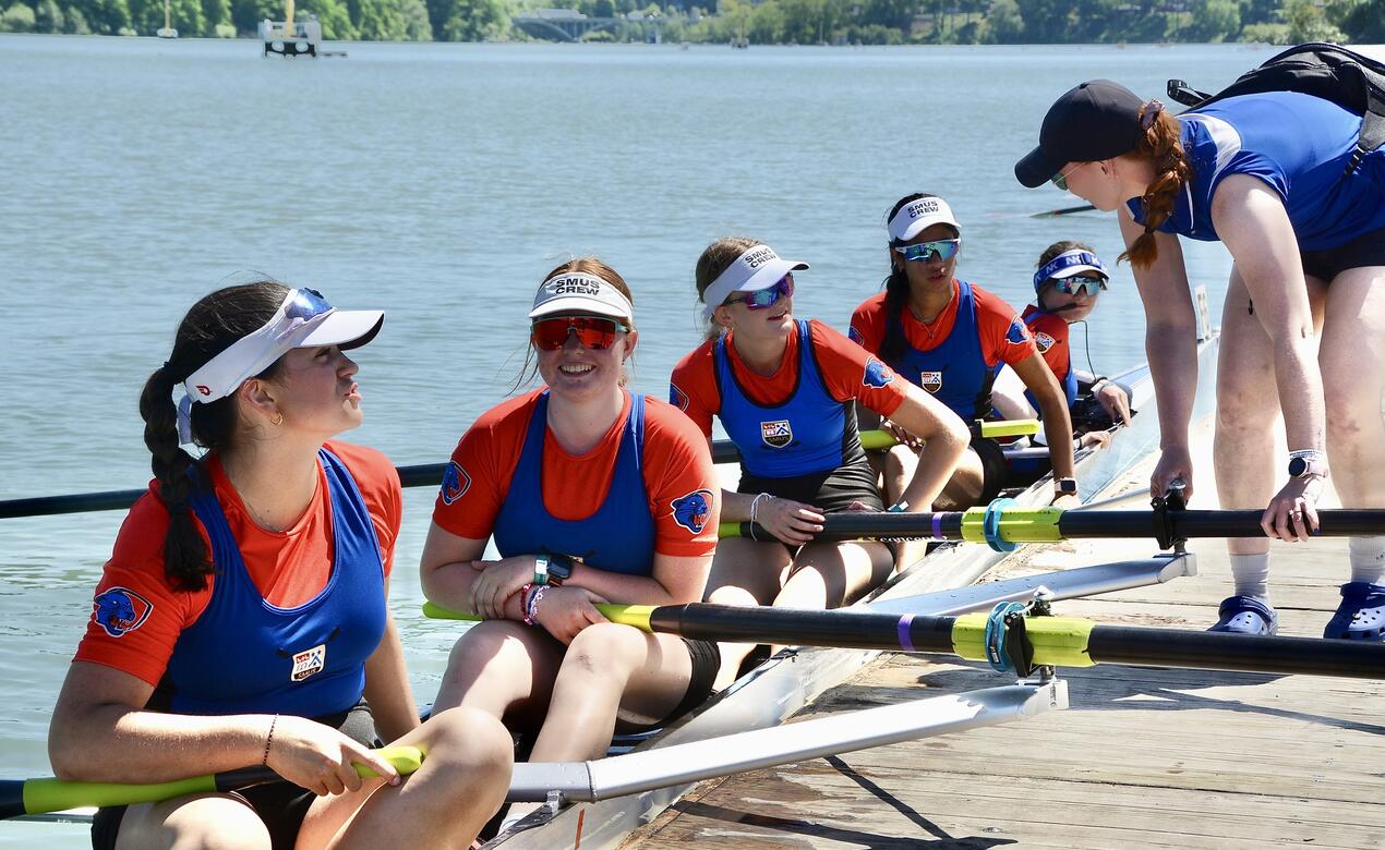 SMUS coxless four women sit in their boat talking to a coach as they prepare for a race. 