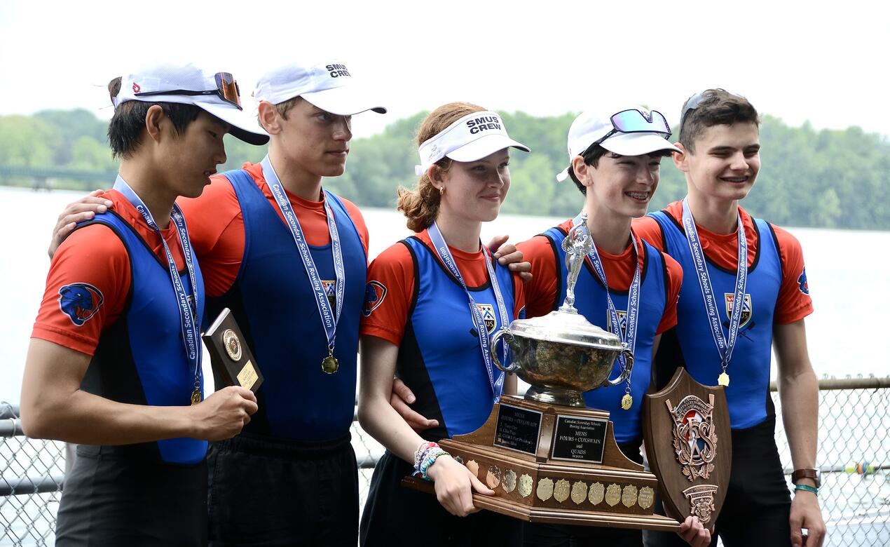 Five SMUS rowers pose for a photo with their medals, plaques and trophy