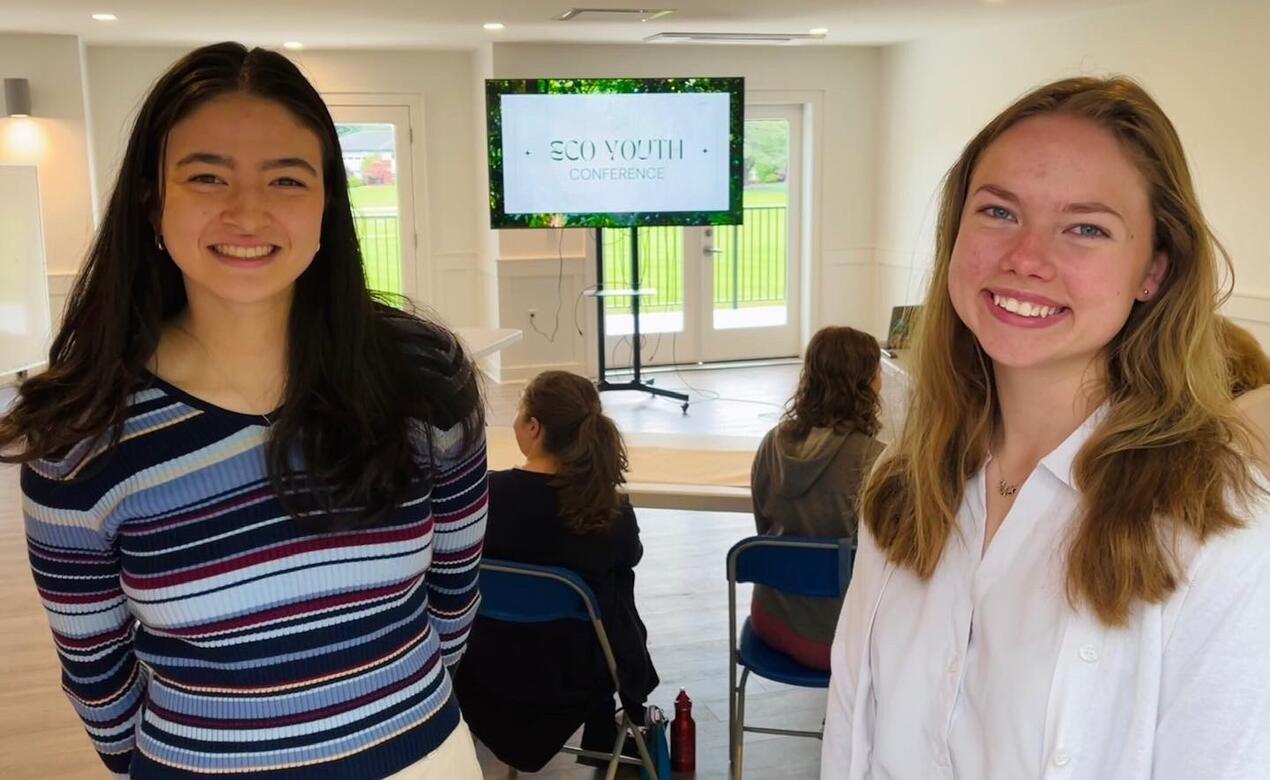 Grade 12's Janet Andrews and Grade 11's Beatrice Sharpe smile at the camera as the first-ever Eco-Youth Conference takes place behind them. 