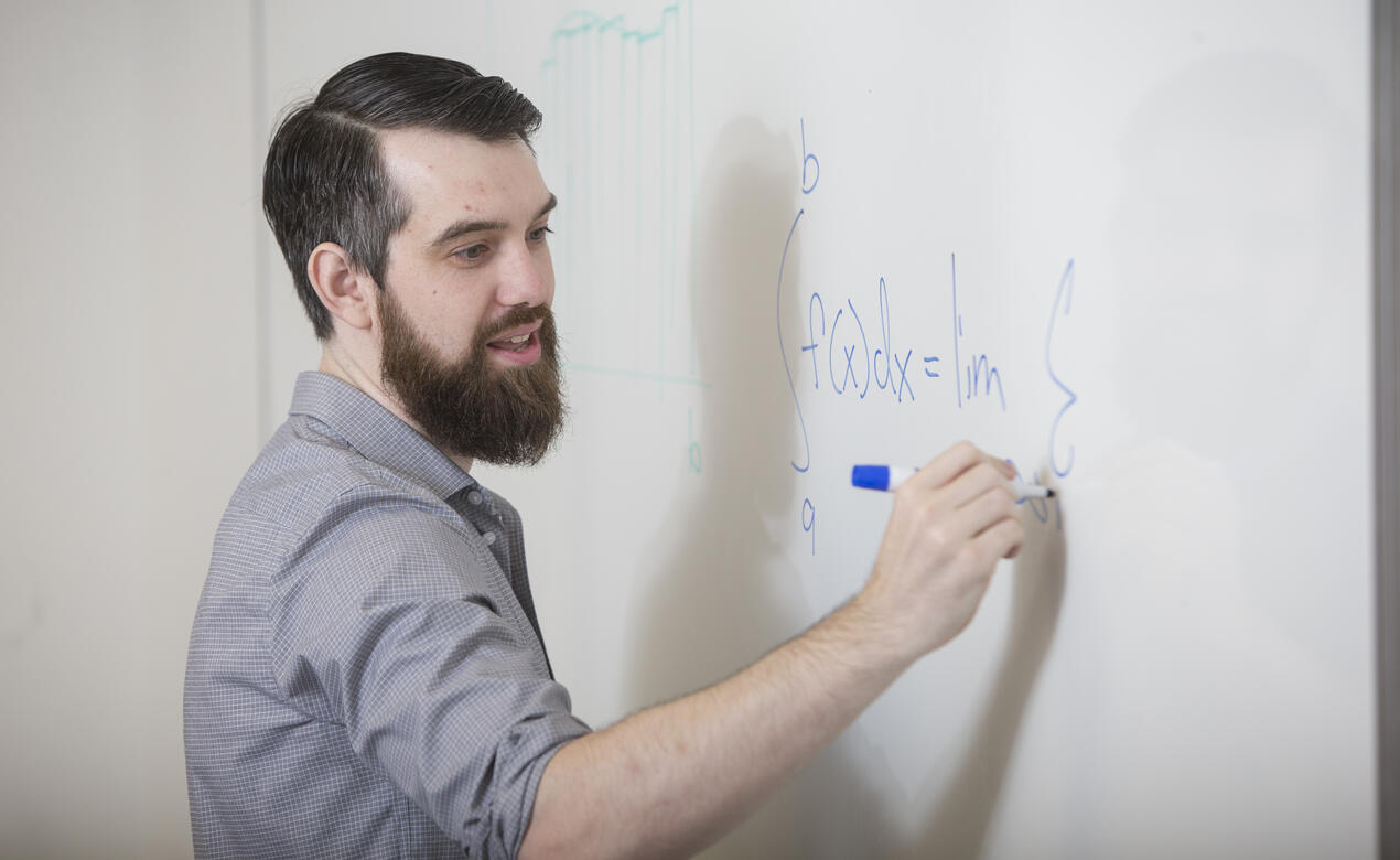 Alumni Dr. Trefor Bazett is writing a math equation on a white board while teaching a class. 