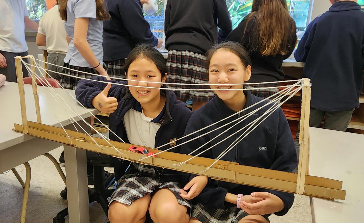 Two students smile as they crouch down in view of their bridge design that connects two desks. 