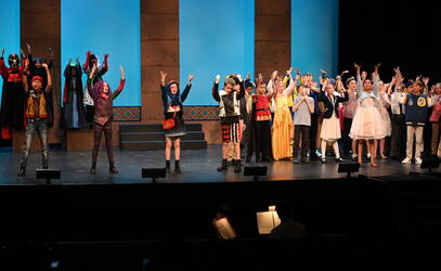A cast of Grade 5 students in costume are across a theatre stage singing with their hands in the air.