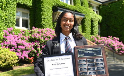 Maya Achuthan stands in front of the SMUS School House Building holding up a certificate and plaque acknowledging her as a Schulich Award nominee.