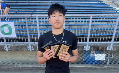 Grade 12 student Leupold Wang stands in front of the stadium stands holding three awards. 