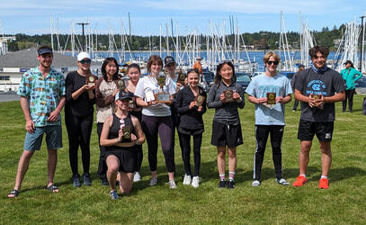 A group photo of the SMUS Sailing team posing with their awards as the harbour of boats are behind them. 