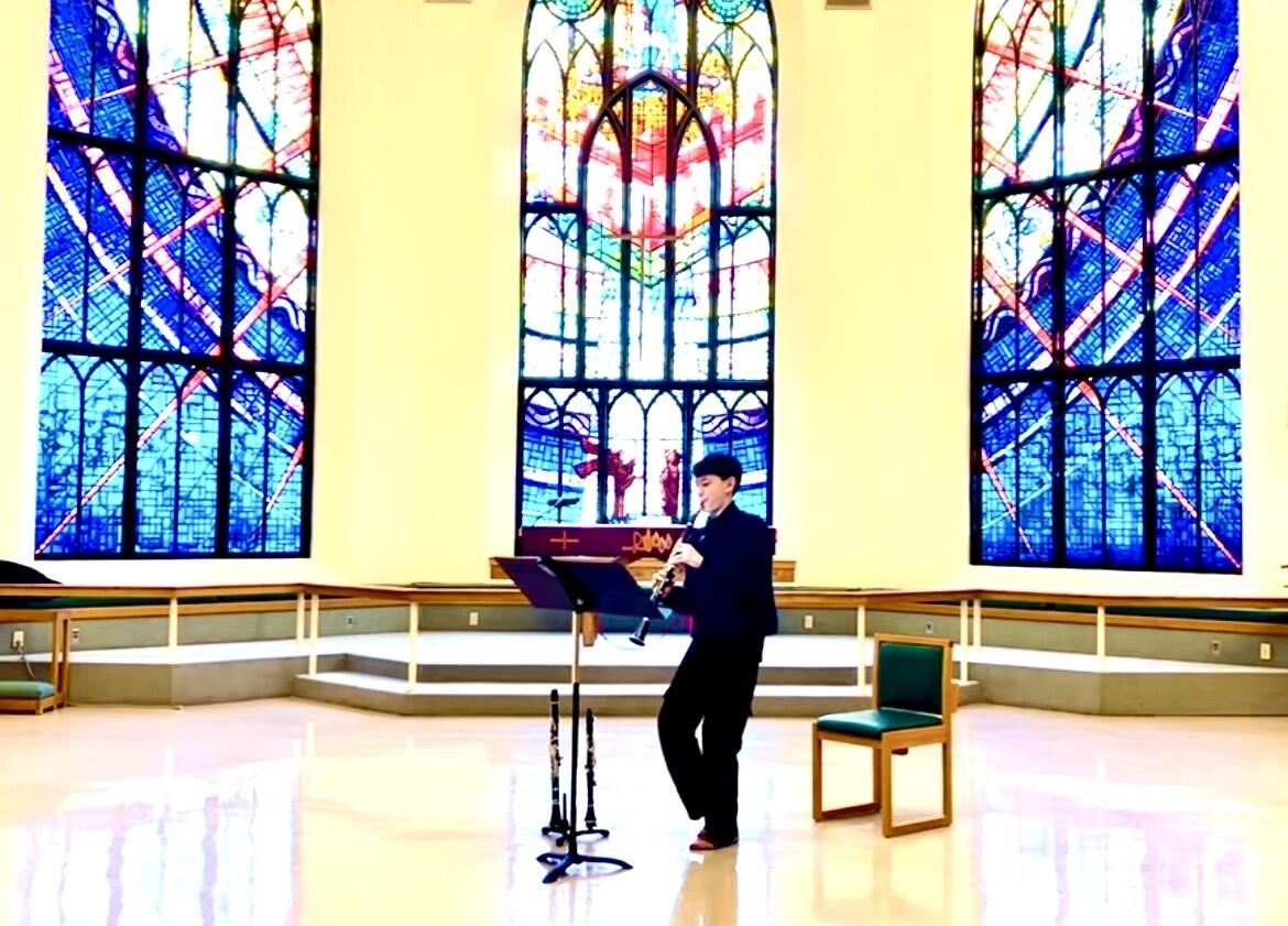 Matty Angus stands inside a church with large stained glass windows while playing clarinet